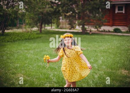 a charming girl in a yellow dress dances with a wreath of yellow flowers on her head on a lawn of green grass outdoors on a sunny spring day Stock Photo
