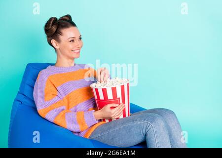Portrait of attractive cheerful girl sitting in chair eating corn watching series having fun isolated over bright teal turquoise color background Stock Photo
