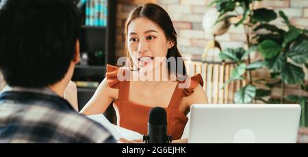 Cheerful young Asian female podcaster holding a questionnaire while interviewing male guest at workspace with mic and laptop Stock Photo