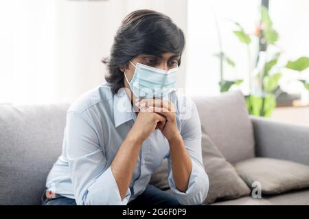 Self Isolation concept. Pensive indian guy wearing protective medical mask sitting on the couch lost in thoughts, staying at home with viral disease symptoms. Pandemic and quarantine concept Stock Photo