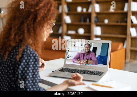 Redhead curly woman sitting at the desk and making video call on the laptop to African-American man, talks with online tutor, learning foreign language with tutor, taking online course Stock Photo