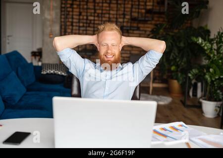 Happy serene redhead businessman in smart casual shirt holding hands behind head rests on the workplace, satisfied with the work done, relaxed freelancer guy takes a break Stock Photo