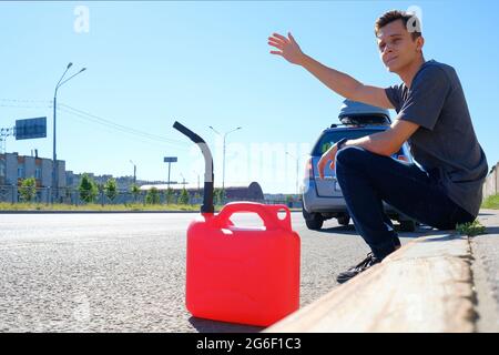 A red canister on the asphalt near the car. The car ran out of gas and stalled. A young man hoping for help on the road from other drivers Stock Photo