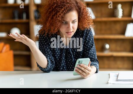 Shocked red-haired curly woman holding a smartphone, feel astonishment with a bad news, fired from work, irritated female eoffice employee looks at phone screen and does not understand what happened Stock Photo
