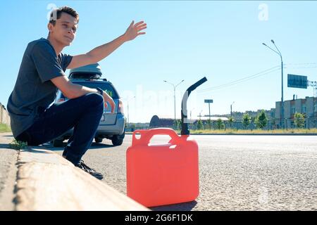 A red canister on the asphalt near the car. The car ran out of gas and stalled. A young man hoping for help on the road from other drivers Stock Photo