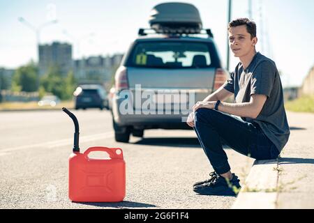 Ran out of gas. A red canister on the asphalt near the car. A young man hoping for help on the road from other drivers Stock Photo