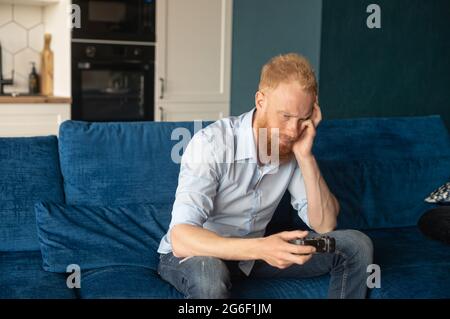 Upset young redhead man loses in video game, sitting on the couch at home with joystick, unhappy and disappointed guy Stock Photo