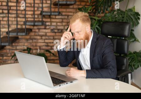 Thoughtful redhead bearded businessman staring at laptop screen and rested head on the hand, solving difficult business tasks, pondering carefully before making a decision or reaching a conclusion Stock Photo