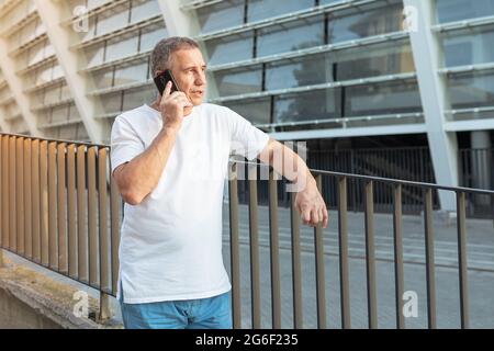 Middle aged man in summer sportswear talking on the phone outdoors on blurred geometric architecture background Stock Photo