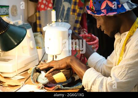 African tailor smiling sewing a hat with sewing machine at sewing workshop. Black man working with african fabrics. Stock Photo