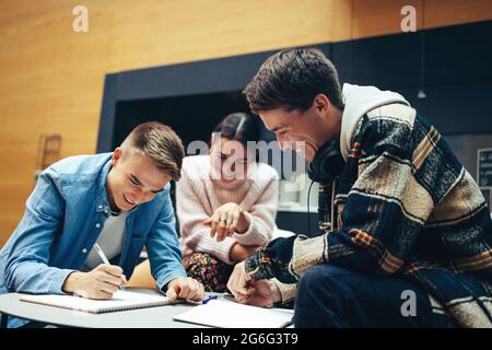 Young man writing notes while studying with friends in high school. Happy students working together on college assignment. Stock Photo
