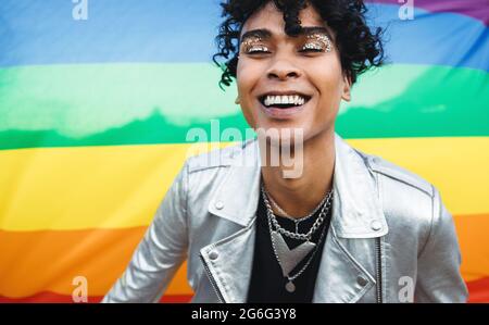 Young gay man embracing his queer identity. Happy gay man smiling cheerfully while standing against a pride flag. Gender non-conforming young man wear Stock Photo