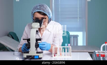 Young woman in glasses and white uniform looks at sample through contemporary microscope sitting at workplace in clinic laboratory Stock Photo