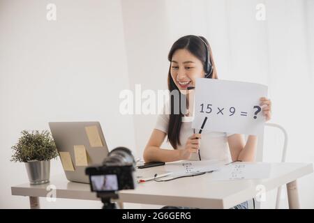 Young beautiful Asian teacher woman teach math online and look at laptop to chat with student in front of camera during quarantine and work from home Stock Photo