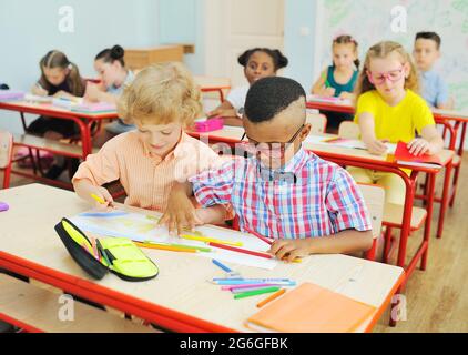 two small children of elementary school students are preparing for a lesson sitting at a Desk with colored pencils in their hands Stock Photo