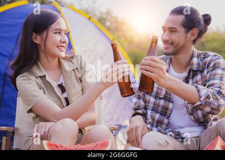 friendship Asian people drinking in camping, beautiful woman holding bottle of beer and cheer with man sitting looking view near tent, freedom and hap Stock Photo