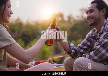 friendship Asian people drinking in camping, beautiful woman holding bottle of beer and cheer with man sitting looking view near tent, freedom and hap Stock Photo