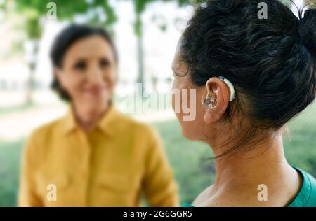 Adult woman with a hearing impairment uses a hearing aid to communicate with her female friend at city park. Hearing solutions Stock Photo