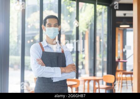 portrait waiter in café, coffee shop staff, smart Asian man wearing mask and cross arm Stock Photo