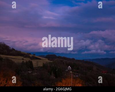 Massive clouds over hilly landscape in winter without snow, with shining street lamp in the foreground and tiny lights in the garden in the background Stock Photo