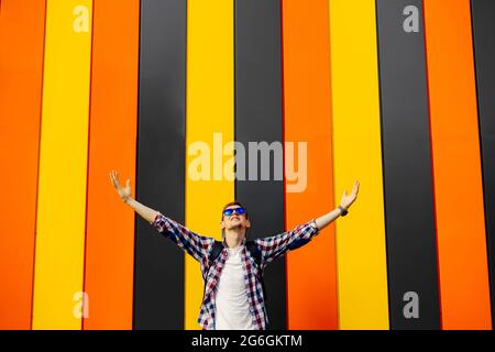 Portrait of happy young man in sunglasses and hat, hipster with raised arms up, excited man with raised arms shouting, on against colorful colorful wa Stock Photo