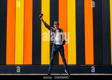 Excited happy young man, in sunglasses and a cap, shows a gesture of success, celebrating victory jumping up with a mobile phone, against the backgrou Stock Photo