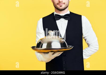 Young handsome waiter holding metal tray with cover ready to serve on yellow background Stock Photo