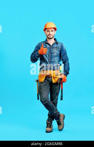 Full length portrait of confident handsome male construction worker in safety helmet showing thumbs up sign over blue background looking to camera Stock Photo