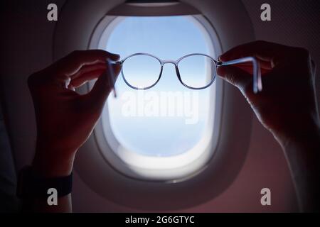 Hand of passenger holding eyeglasses against airplane window during flight. Themes travel, eyesight and vision. Stock Photo