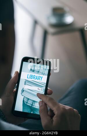 a young man, sitting at a coffee table in his living room, is searching an ebook to lend on an online library with his smartphone, with a simulated se Stock Photo