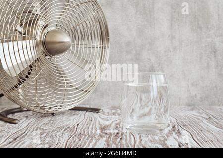 Transparent glass of clear water and a small table fan on a wooden table closeup, shallow depth of field Stock Photo