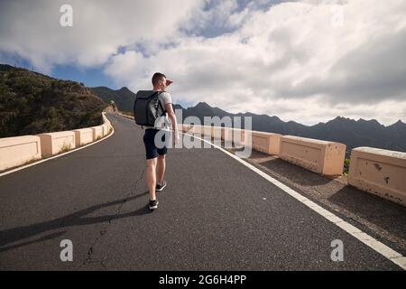 Rear view of man with backpack on mountain road. Young tourist enjoys trip on sunny day. Tenerife, Canary Islands, Spain. Stock Photo