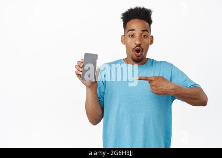 Image of thrilled african american guy pointing finger at smartphone screen, empty mobile phone display, gasp amazed and say wow, checking out new Stock Photo