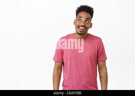 Happy african american man smiling thoughtful, looking at upper left corner and imaging smth tasty, dreaming of delicious thing, standing in pink t Stock Photo