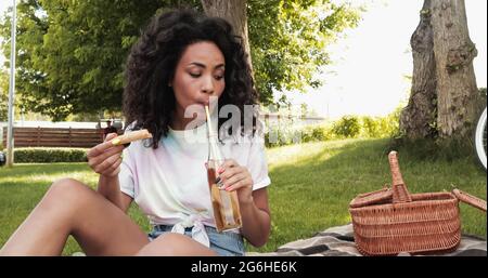 african american woman drinking lemonade through straw and holding piece of pizza during picnic Stock Photo