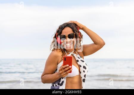 Happy smiling woman with red headphones and smartphone on beach landscape.Female using phone listening music on the seaside. Stock Photo