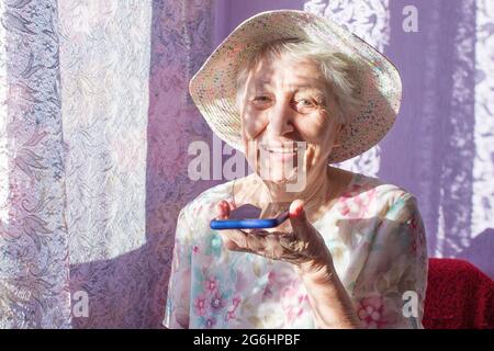 Charming elderly woman in rectangular black rimmed eyewear using voice recognition function on cell phone. Modern senior female holding electronic gadget, sending vocal message via online messenger Stock Photo