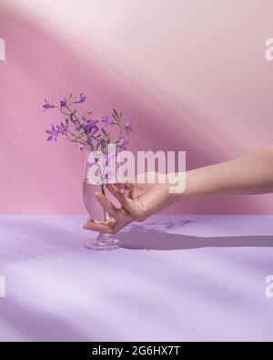 Premium Photo  Hand holding glass with transparent liquid and beautiful  purple field flowers. pastel pink summer or spring background. minimal  nature, aesthetic. natural sun rays and shadows.