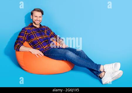 Full size profile portrait of satisfied young man sit on big soft chair beaming smile isolated on blue color background Stock Photo
