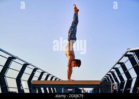 Fit man doing a hand stand while practicing yoga alone near the ocean against sky at dusk or dawn Stock Photo