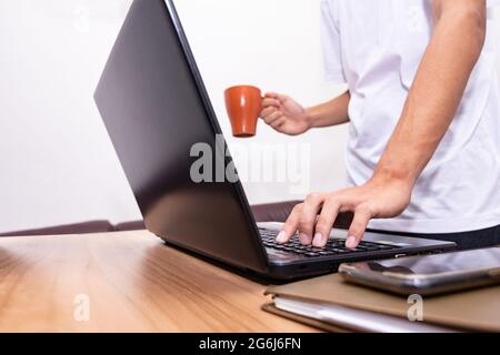 The businessman is standing holding a coffee cup and working on a laptop at his home. technology, people, and lifestyle concept. Stock Photo