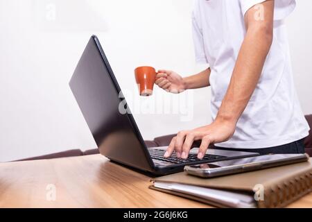 The businessman is standing holding a coffee cup and working on a laptop at his home. technology, people, and lifestyle concept. Stock Photo