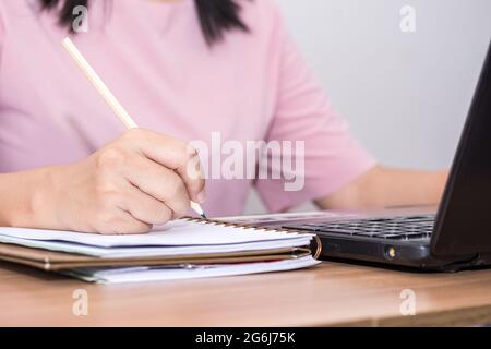 Closeup a woman hand holding pencil and using a laptop computer working at her desk in the office. A businesswoman is writing down her work in a noteb Stock Photo