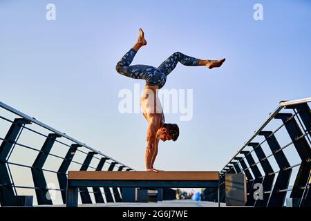Fit man doing a hand stand while practicing yoga alone near the ocean against sky at dusk or dawn Stock Photo