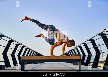 Fit man doing a hand stand while practicing yoga alone near the ocean against sky at dusk or dawn Stock Photo