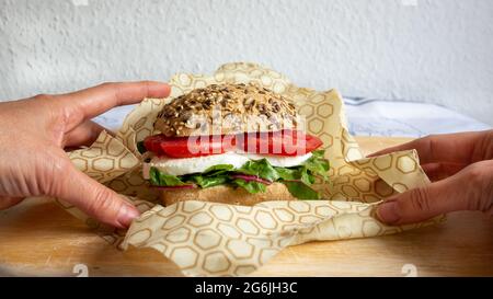 Two hands packing with bee wax recyclable wrapping  a lunch sandwich with bread bun, mozzarella cheese and lettuce. Zero waste. Close up. Stock Photo