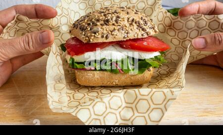 Two hands packing with bee wax recyclable wrapping a lunch sandwich with bread bun, mozzarella cheese and lettuce. Sustainable packaging. Keep fresh. Stock Photo
