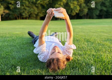 Guy teenager lying on grass with smartphone. Male in white T-shirt with phone in his hands, top view, green lawn background Stock Photo