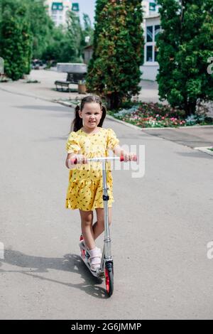 full-length portrait of a charming girl in a yellow summer dress riding a scooter on an asphalt path along an alley with cypresses Stock Photo