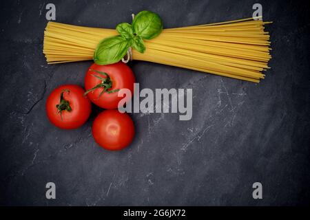 A raw bunch of pasta garnished with a sprig of basil and three fresh tomatoes on a black slate plate Stock Photo